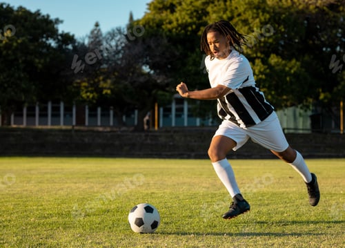 Preview: Male soccer player running toward ball on sunlit field, with striped jersey, cleats, copy space