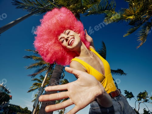 Preview: Bright pinkhaired woman extends hand toward a palm tree, standing in an outdoor tropical setting