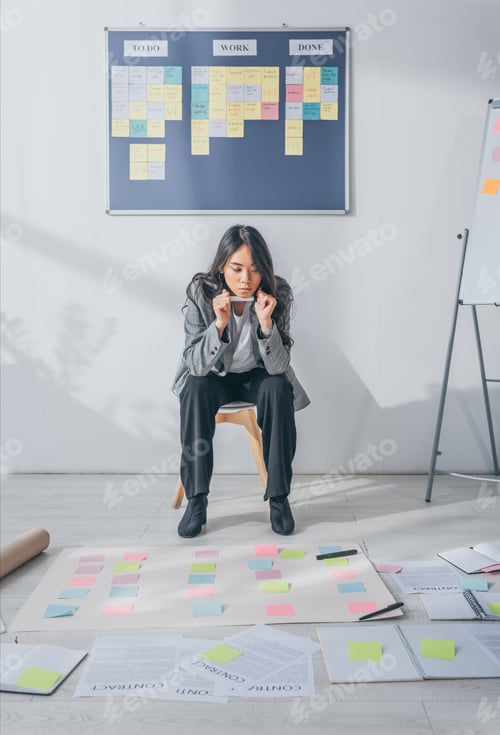 Preview: pensive asian scrum master sitting on chair and looking at sticky notes