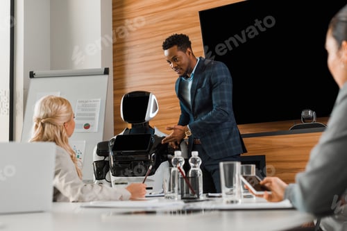 Preview: handsome african american businessman pointing with finger at robot near colleagues in conference