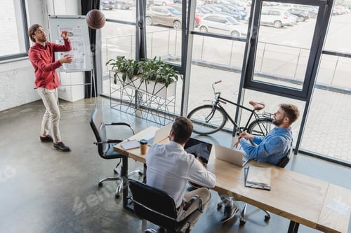 Preview: high angle view of young businessmen playing with basketball ball and using laptops in office