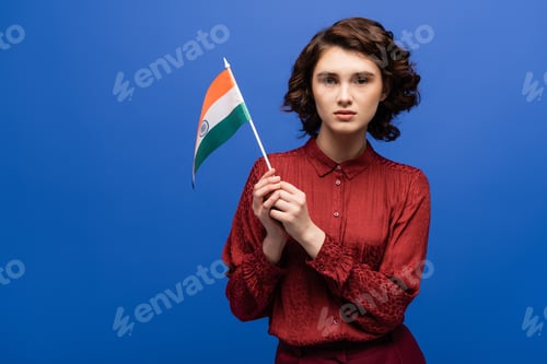 Preview: curly language teacher holding flag of India and looking at camera isolated on blue