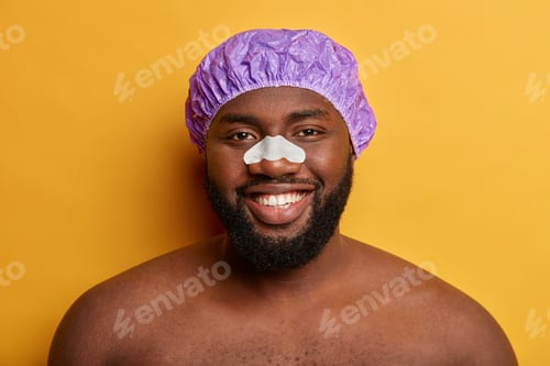 Preview: Handsome dark skinned man uses nose patch for reducing black dots and wrinkles, wears shower cap, sm