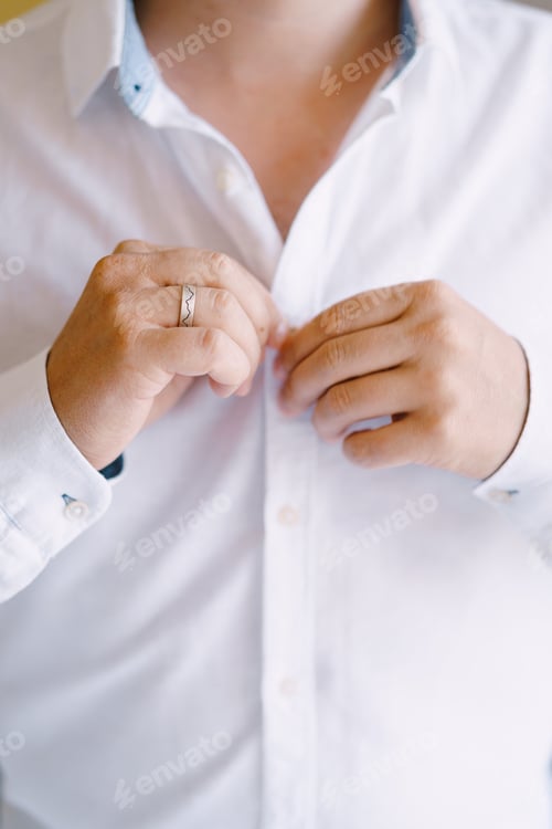 Preview: Groom is buttoning the buttons on the white shirt. Close-up