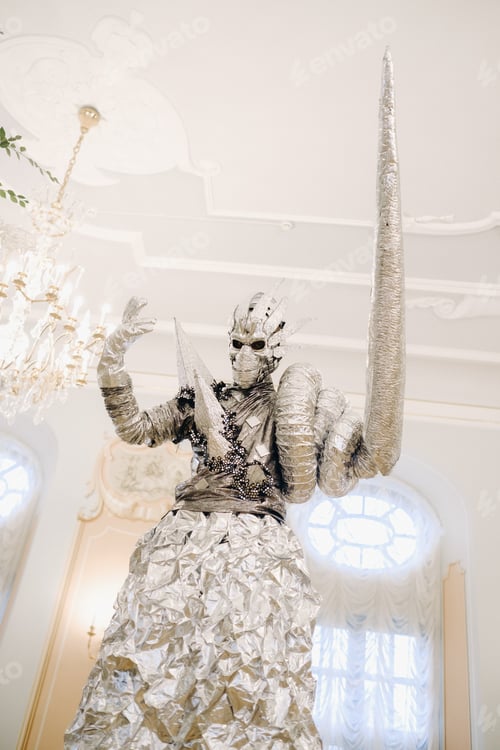 Preview: A man in a silver suit and a mask on stilts in the interior of the castle