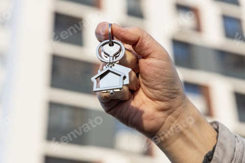 Preview: Keychain in house shape and key in male hand on background of new buildings