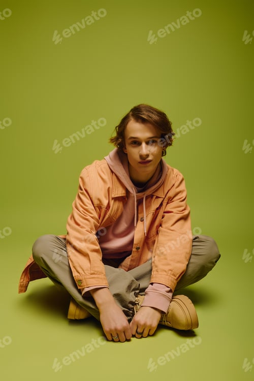 Preview: Young man exudes modern style while sitting on a vibrant green backdrop with utmost confidence