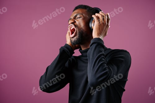 Preview: A young Black man expressing strong emotions while wearing headphones against a pink background