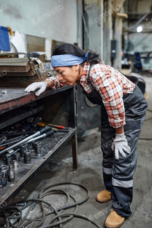 Preview: Young female technician in workwear bending in front of workbench with tools
