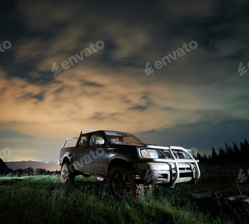 Preview: Illuminated pickup offroad truck in mud on mountain meadow at night under starry cloudy sky
