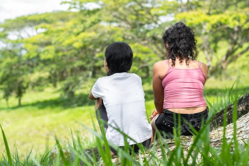 Preview: two brunette latina girls, sitting next to a tree in a tall grass field admiring nature