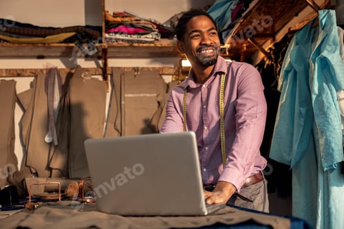 Preview: Male fashion tailor having video call with customer via laptop for the suit design in his workshop.
