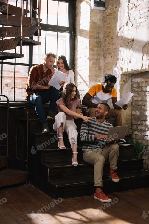 Preview: young, smiling multicultural businesspeople talking while sitting on stairs