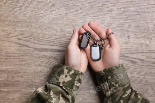 Preview: Man in camouflage uniform holding military ID tags over wooden background, top view