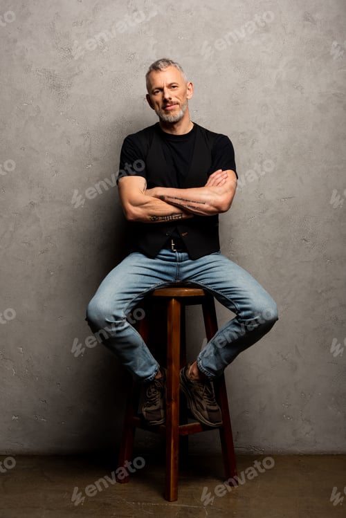 Preview: bearded man with crossed arms sitting on stool on grey