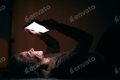 Preview: Smiling Woman With Face Illuminated By Digital Tablet Screen Lying On Carpet At Night