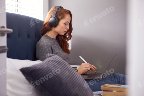 Preview: Female College Student Wearing Wireless Headphones Sitting On Bed In Shared House Using Laptop