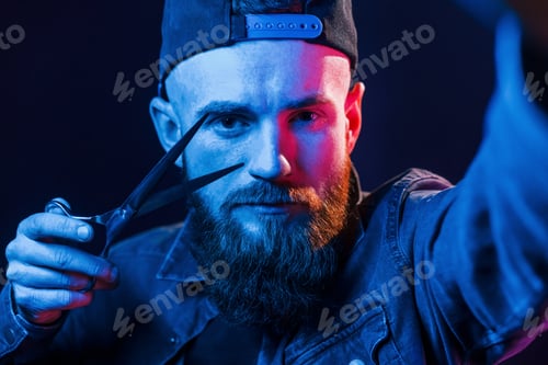 Preview: With scissors. Young bearded barber in cap standing in the studio with neon lighting