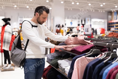 Preview: Adult handsome brunet man in white sweatshirt chooses stylish trendy shirt at clothing store in