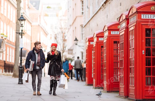 Preview: Young shopping couple strolling past red phone boxes, London, UK