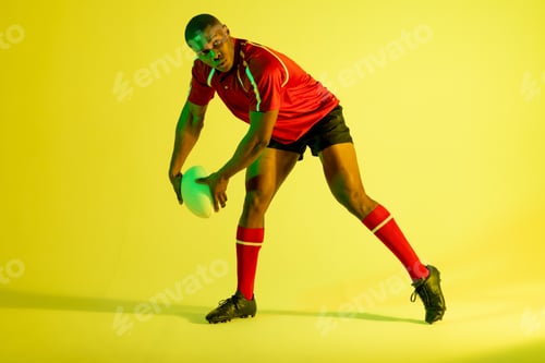 Preview: African american male rugby player with rugby ball over yellow lighting