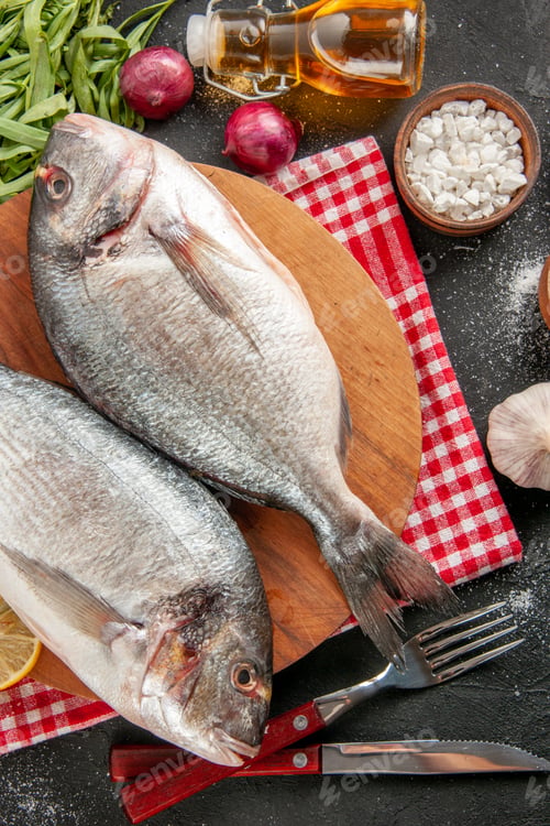 Preview: top view fresh raw fish on round wood board sea salt fork and knife tarragon garlic lemon slices oil