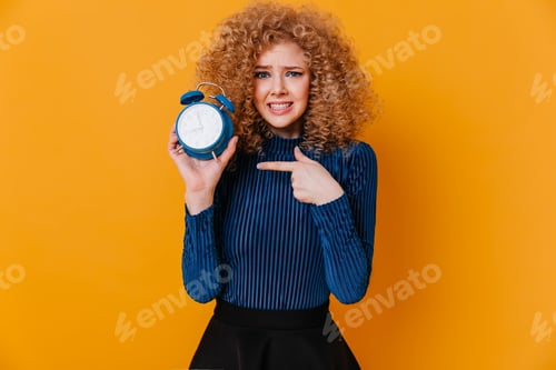 Preview: Girl feels uncomfortable with ringing alarm clock. Portrait of curly woman in blue blouse on yellow