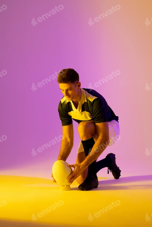 Preview: Caucasian male rugby player crouching with rugby ball over pink lighting