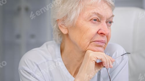Preview: Thoughtful aged lady with sad eyes wrinkly face alooks aside holding glasses in hand