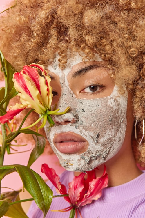 Preview: Vertical shot of curly haired young woman applies beauty clay mask covers face with exotic plant und