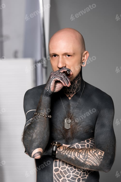 Preview: bald shirtless man with tattoos smoking cigar and looking at camera on grey
