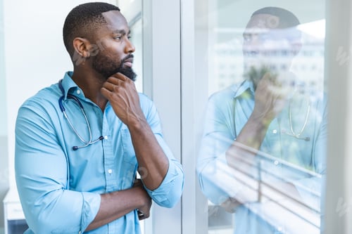Preview: Thoughtful african american male doctor wearing blue shirt looking through window in hospital room