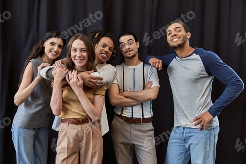 Preview: joyful multiethnic colleagues embracing and looking at camera on black backdrop in modern office