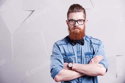 Preview: Looking trendy. Handsome young bearded man in glasses keeping arms crossed and looking at camera