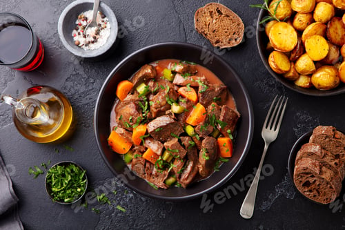 Preview: Beef Meat and Vegetables Stew in Black Bowl with Roasted Baby Potatoes. Dark Background. Top View.