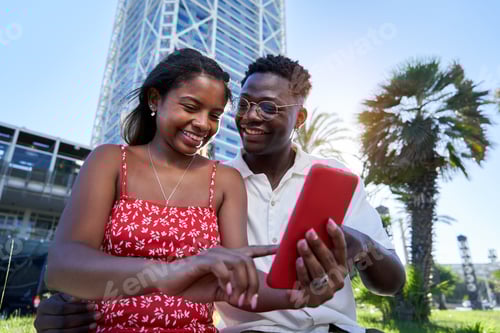 Preview: Young African American couple sitting outdoors using smart phone together on summer holidays.