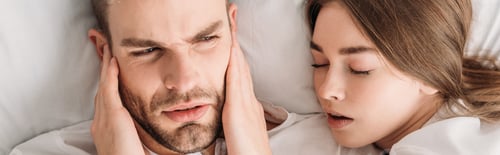 Preview: top view of exhausted man plugging ears with hands while lying in bed near snoring wife, panoramic