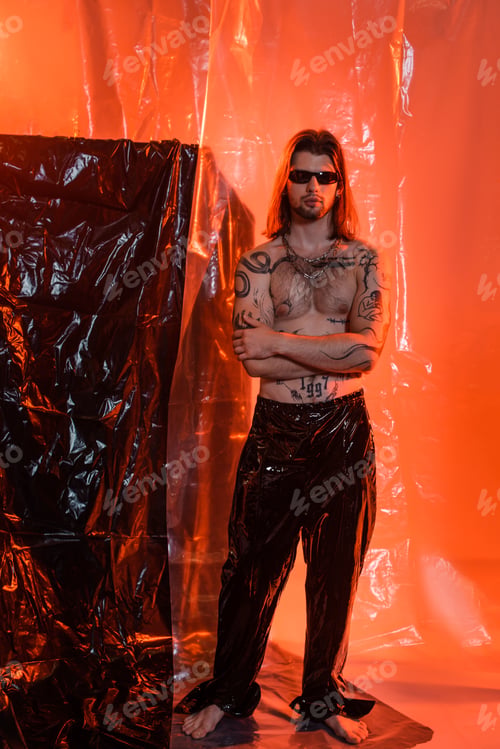 Preview: Tattooed man in latex pants and sunglasses standing near cellophane