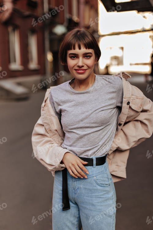 Preview: Short-haired woman smiling outdoors. Happy girl with brunette hair in jeans with belt, t-shirt and