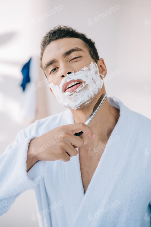 Preview: portrait shot of handsome man in bathrobe shaving face with razor blade and looking at camera