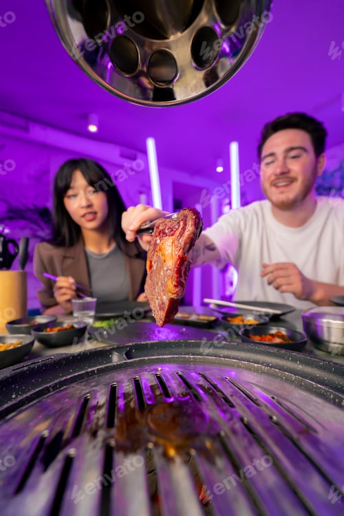 Preview: hand with tongs picking up a juicy raw piece of meat to put on the grill in a Korean restaurant