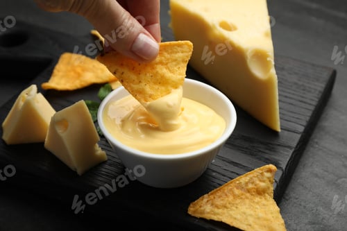 Preview: Woman dipping nacho chip into tasty cheese sauce at black table, closeup