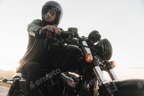 Preview: Handsome biker sitting on his motor bike, wearing leather jacket, helmet and sunglasses