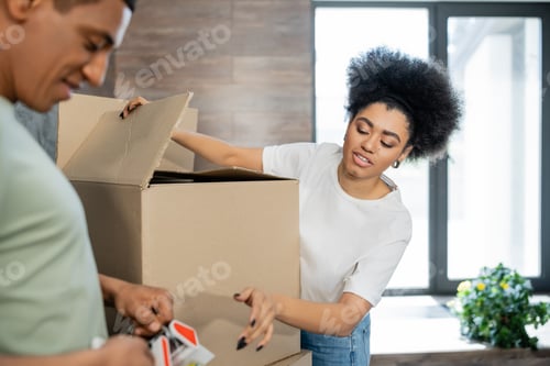 Preview: african american woman talking to boyfriend with adhesive tape near carton boxes during moving