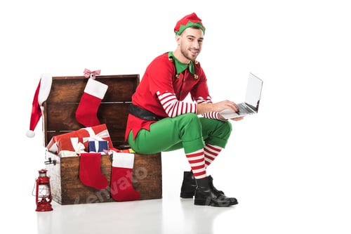 Preview: smiling man in christmas elf costume sitting on wooden box and using laptop isolated on white
