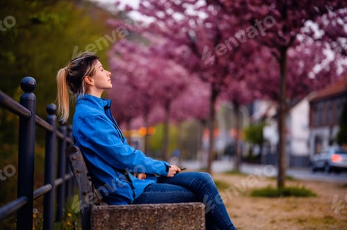 Preview: Young girl in blue jacket and jeans sitting on bench on street with sakura alley resting