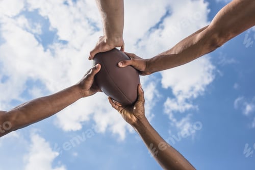 Preview: human hands holding american football ball on background of blue sky