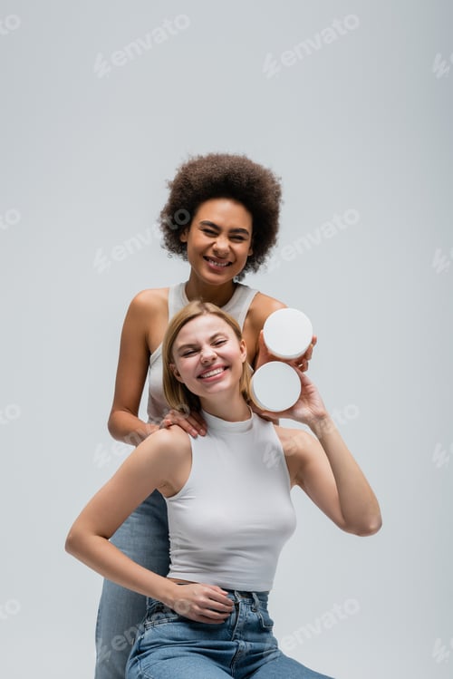 Preview: excited multiethnic models in white tops and jeans posing with body cream and looking at camera