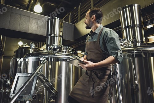 Preview: Male worker in apron inspecting industrial equipment at the brewery