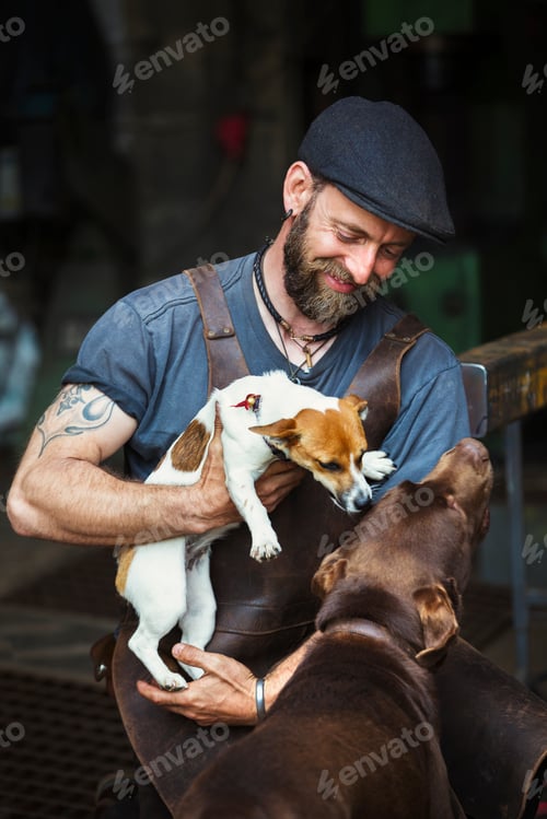 Preview: A man in a leather apron playing with two dogs in a workshop.